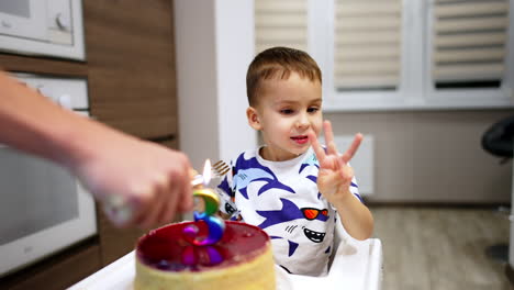 beautiful baby boy sitting in high chair with a fork in hand. parent lights a candle in the shape of number three in front of the kid. happy toddler blows the candle.