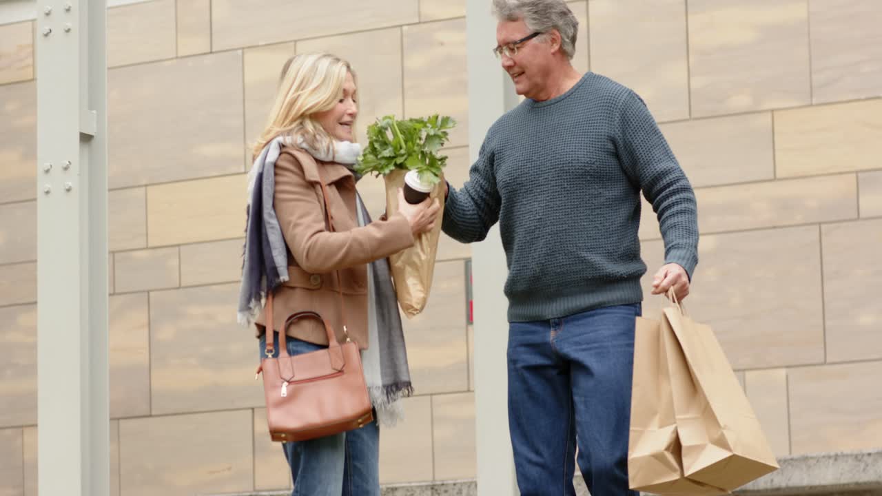 Casal de idosos caminhando no pátio, maçã caindo, homem compartilhando sacola enquanto mulher segura uma planta e uma xícara de café.