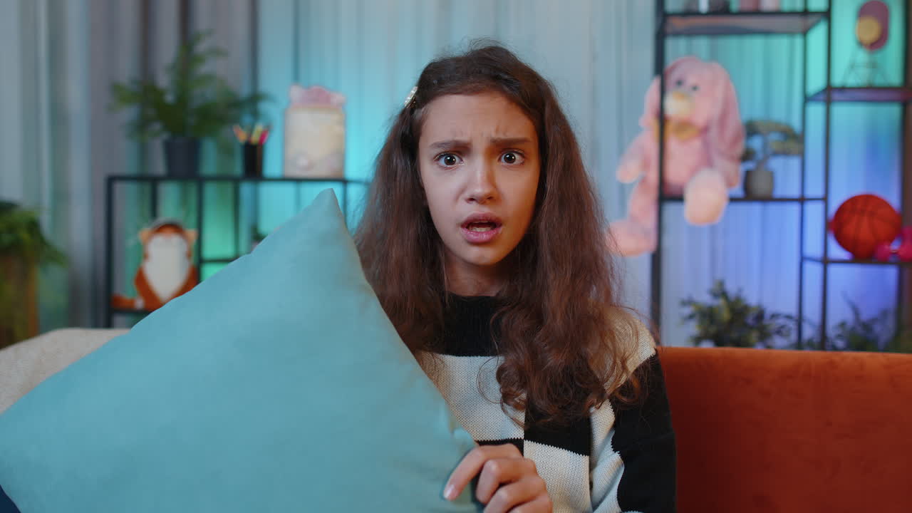 A young girl looking scared and holding a pillow while sitting on a couch