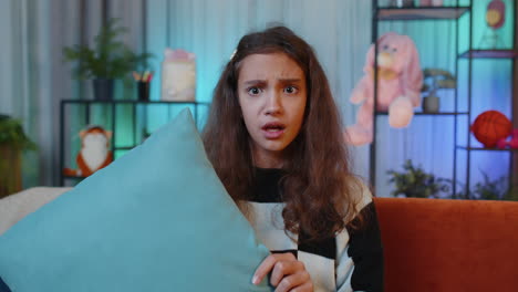 a young girl looking scared and holding a pillow while sitting on a couch