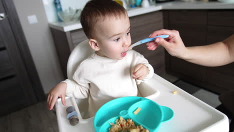 mom shoves a full spoon to her son's mouth. adorable kid takes a spoon from a table. close up. kitchen backdrop.