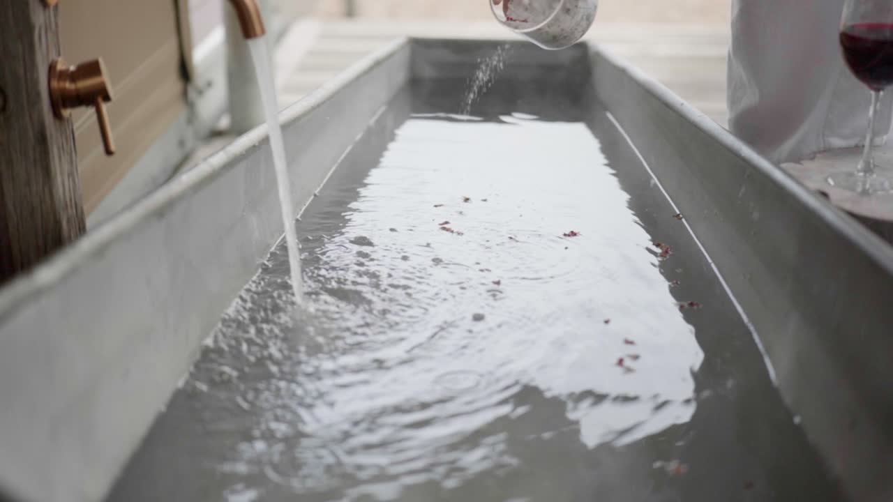 Slow motion close-up of hand pouring bath salts into metal bathtub with water splashing in luxury spa setting with wine glass