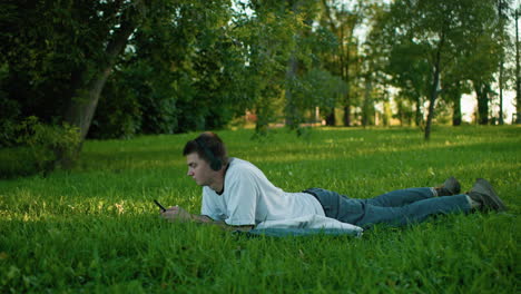 side view of young man lying on mat on grassy field wearing headphones, focused on his phone, nodding his head to music in peaceful green park surrounded by trees under warm sunlight