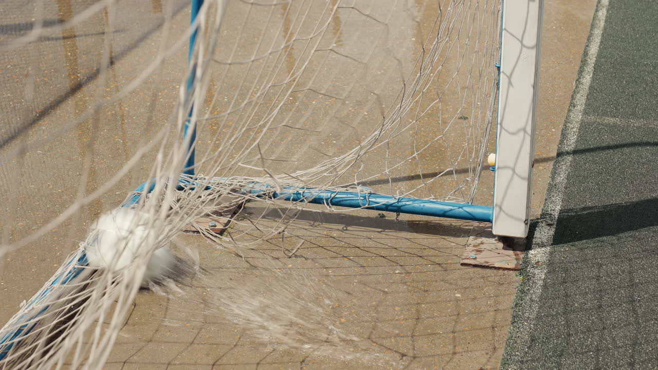 Un pallone da calcio colpisce la rete su un campo bagnato, gocce di pioggia che schizzano intorno all'angolo della porta, primo piano che cattura l'impatto e il rimbalzo, palo blu e rete aggrovigliata visibili, ombra dello stadio vuoto sul campo, trionfante