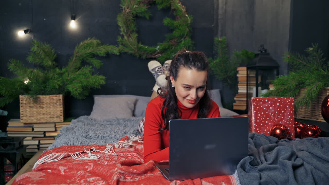 Attractive girl with a laptop on bed. Beautiful young woman looking into the laptop while lying on bed with presents near the new year tree.