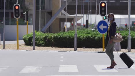 African-american-woman-wearing-face-mask-crossing-road-carrying-suitcase