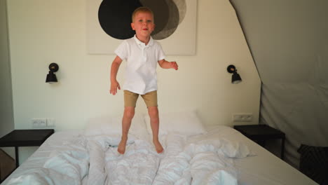 boy stands on bed with snowwhite linen preparing to jump in bedroom of modern apartment. moments from positive childhood. joy of active movement