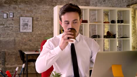 joven empresario feliz está trabajando con una computadora portátil y bebiendo café en la oficina, mirando a la cámara, concepto de negocio, concepto de comunicación
