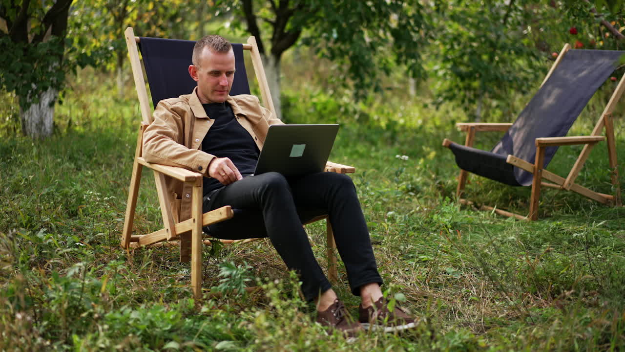 Calm relaxed freelancing man sitting in apple garden with laptop on his knees. Caucasian male working remotely online from nature.