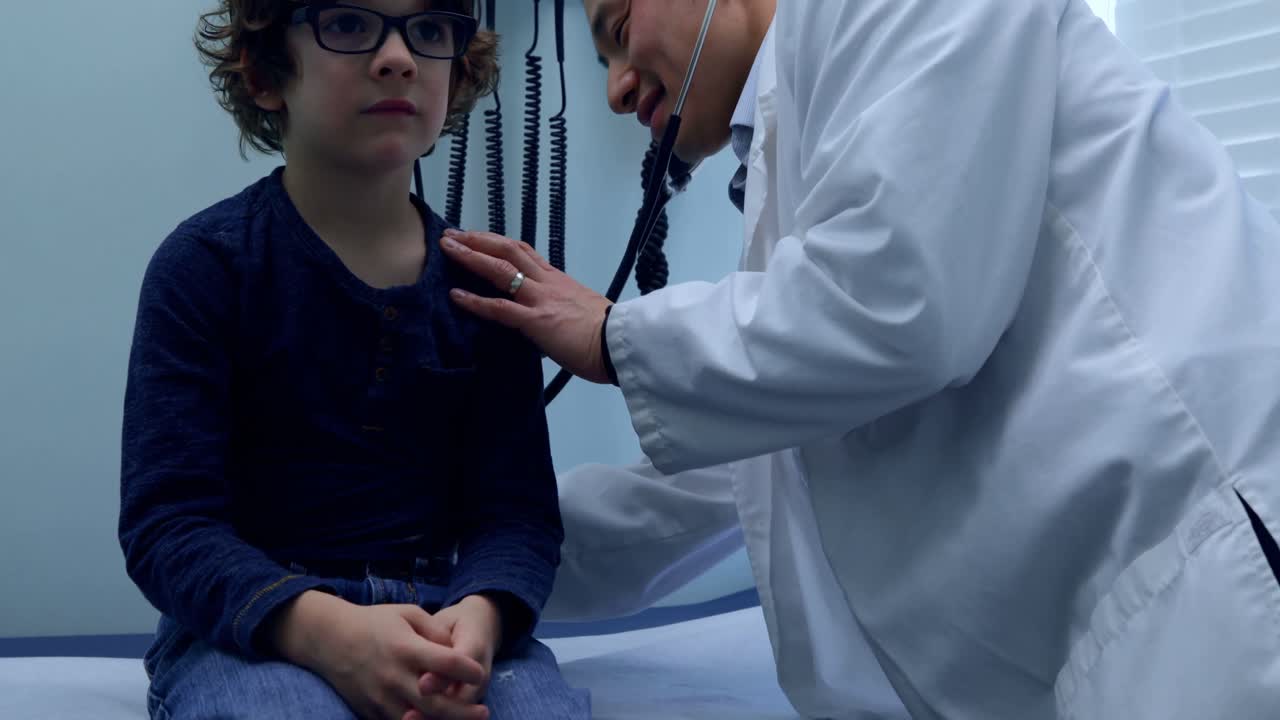 Side view of young asian male doctor examining caucasian boy patient chest in a clinic 4k
