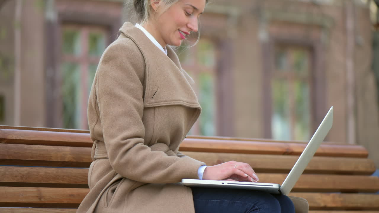 Woman in a brown coat working on her laptop on a bench in the park