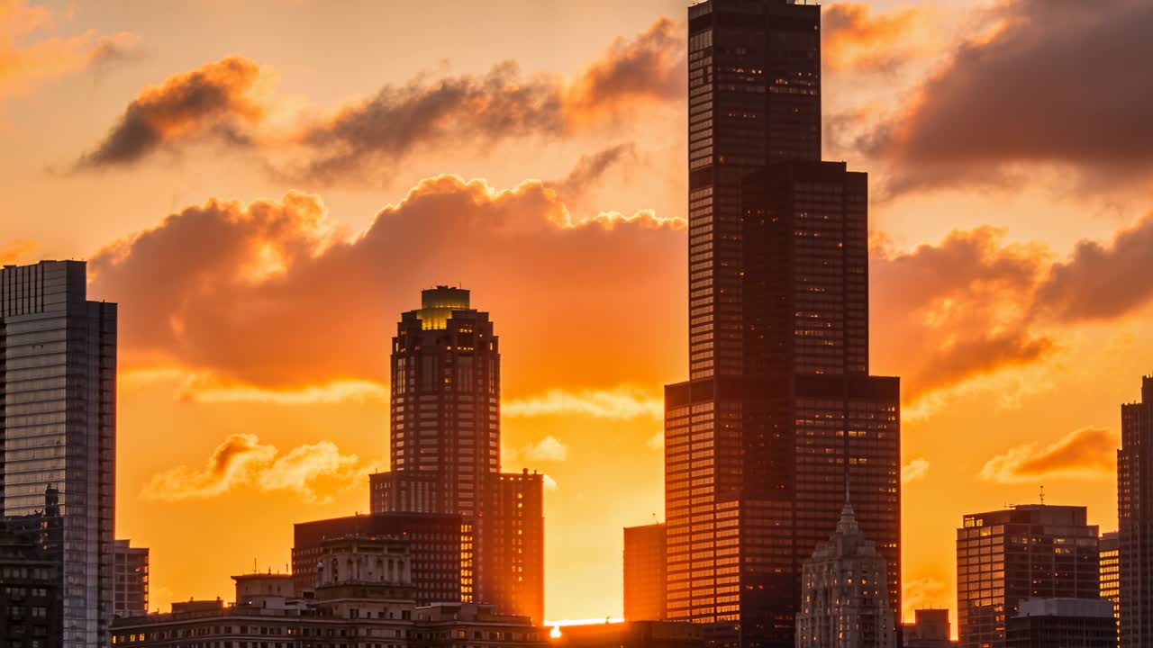 O sol se pondo movendo-se atrás de um aglomerado de torres no centro, banhando a torre mais alta e as nuvens em tons de laranja.