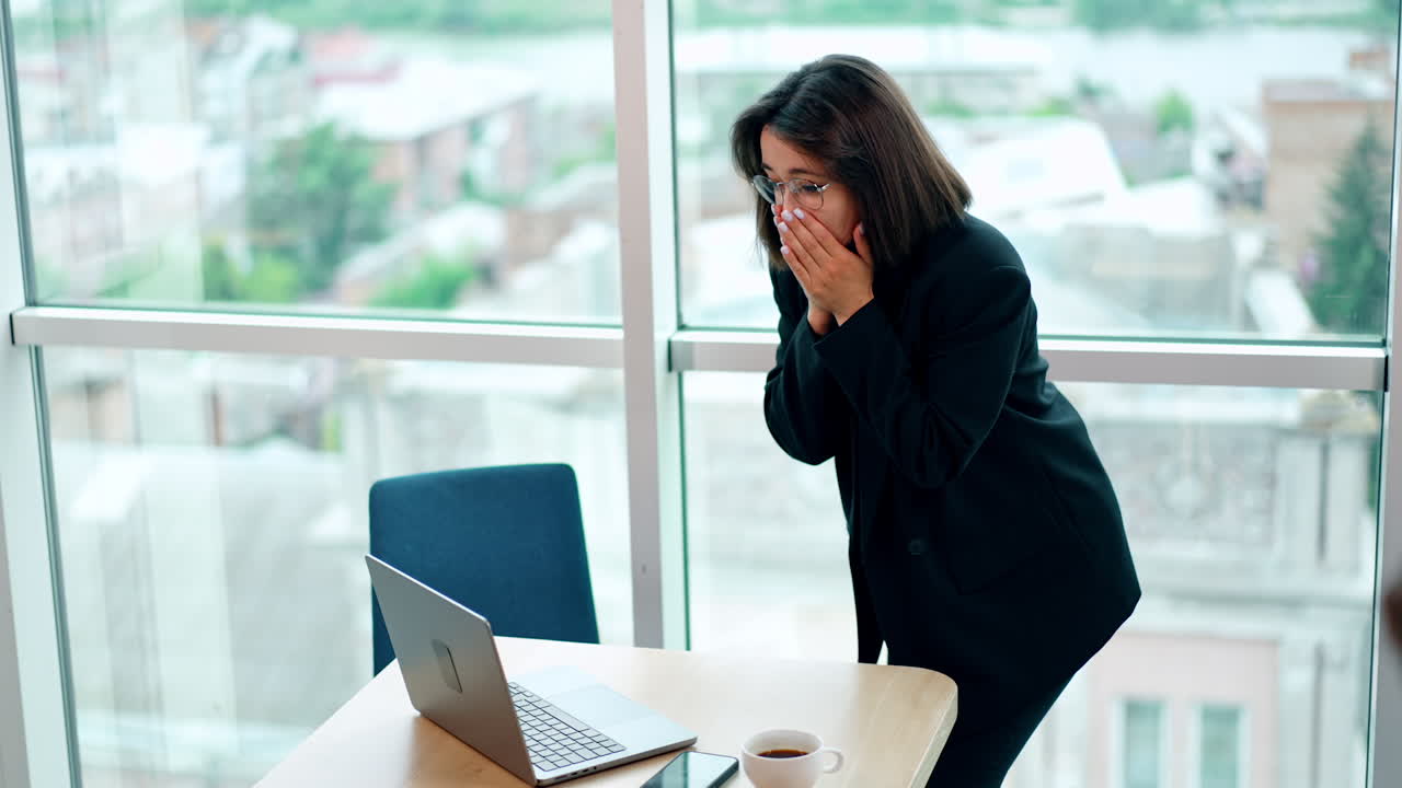 Cheering Caucasian lady leaning to her laptop standing on the desk. Woman receives the good news and jumps and claps with happiness.