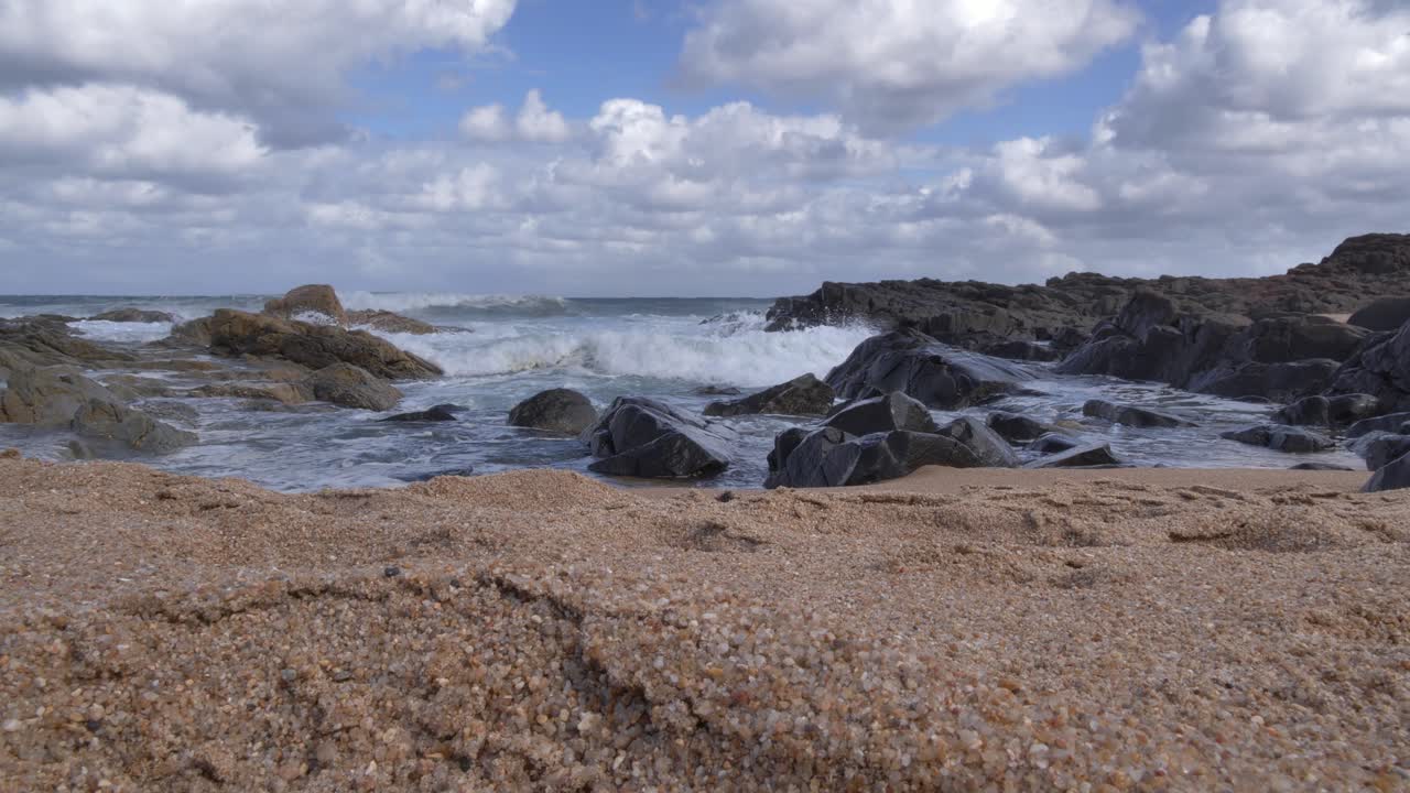 Scenen bevæger sig fra lys til skygge, mens havbølger skyller ind og bryder mod mørke, barske klipper og en tekstureret sandstrand under en dramatisk himmel fyldt med skyer ved Ramsgate, Sydkysten, KwaZulu-Natal