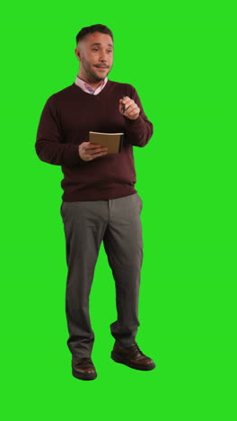 man in a sweater standing in front of a green screen talking and writing in a notebook