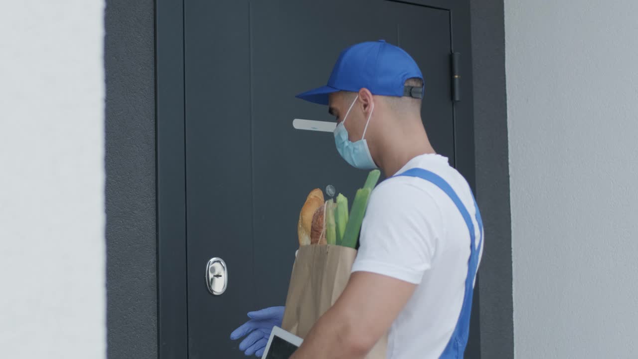 joven mensajero con máscara de protección y guantes entrega un paquete de compras con alimentos directamente a casa de un cliente con seguridad. entrega de alimentos, entrega a casa en cuarentena