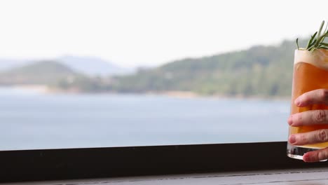 a hand picks up a cocktail from a ledge overlooking a serene seascape, with natural lighting and a calm atmosphere