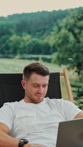 work in the nature far away from the office. relaxed happy freelancing man working on his laptop sitting in a folding chair. vertical video
