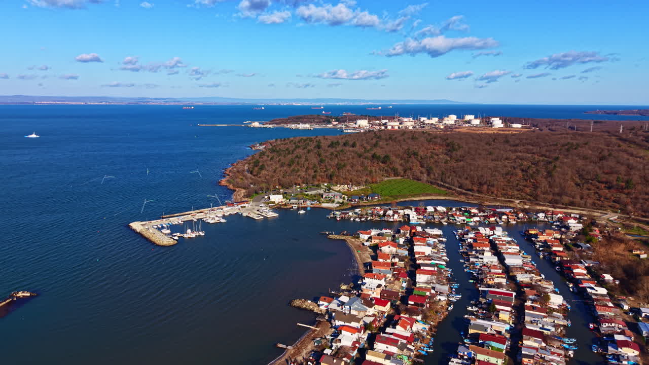Una vista di una città costiera con case lungo la riva e barche ormeggiate al porto turistico in una giornata limpida con cieli azzurri e lontani edifici industriali