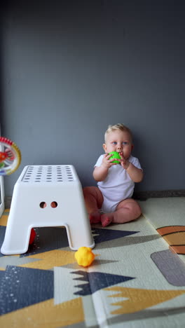 lovely blond baby sitting on the floor near the wall. kid picks a ball from the carpet and takes it to his mouth. vertical video.