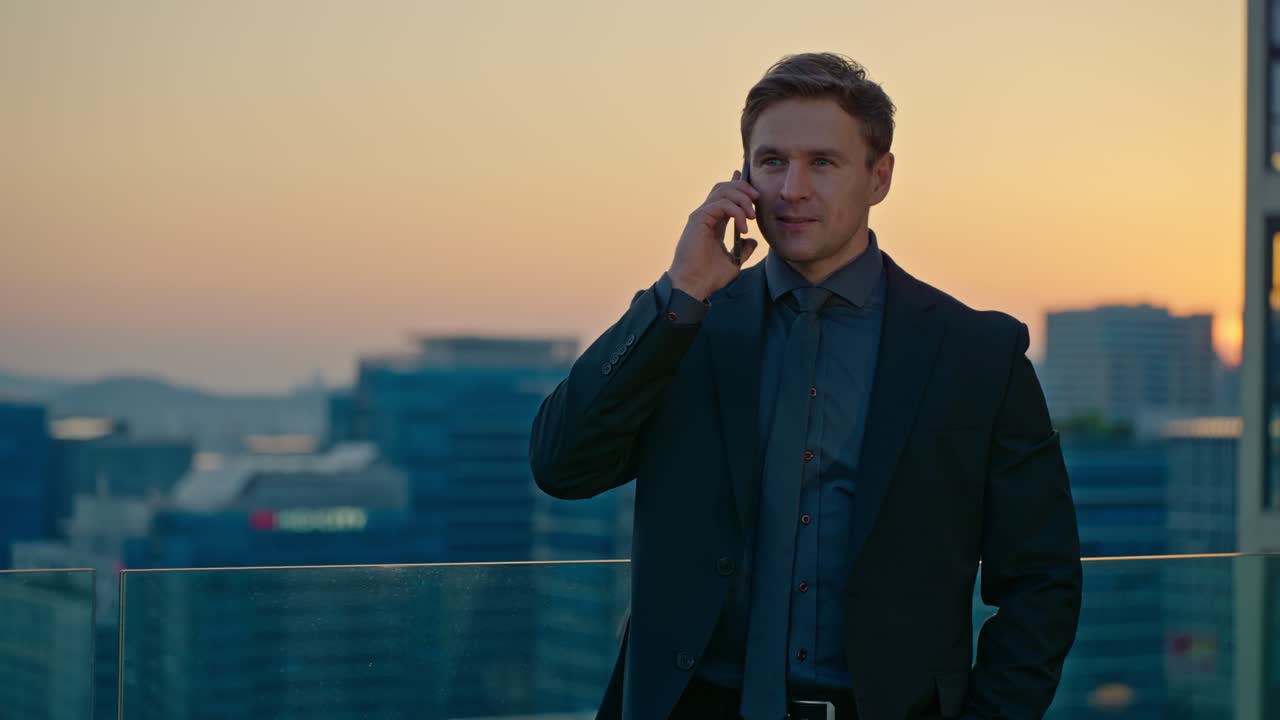 A Caucasian joyful businessman has a pleasing and calm conversation on his smartphone while on a rooftop overlooking the Seoul skyline at sunset