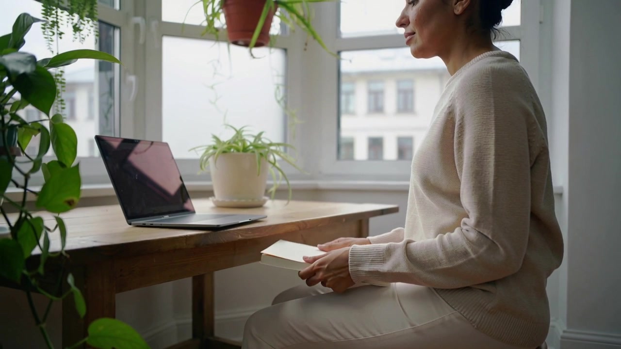 Shifting, woman in knit sweater glancing at laptop at window, lowering notebook to rest, copy space