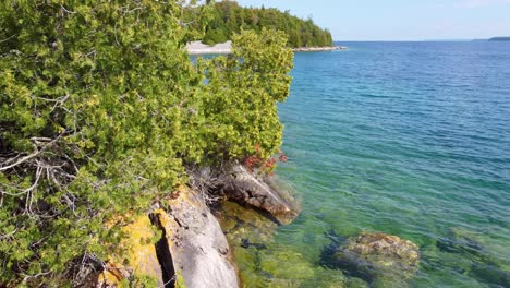 Drone-flies-along-crystal-clear-coastal-water-as-lush-vibrant-green-trees-poke-out-over-rocky-granite-blocks