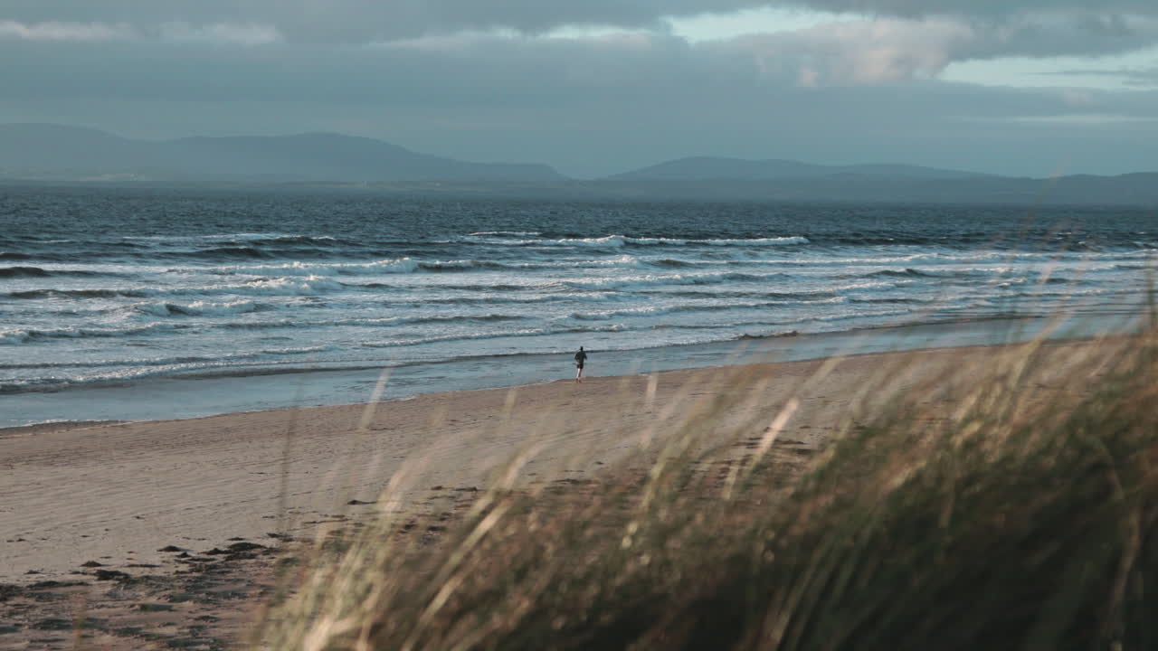 A person strolls along the tranquil beach of County Donegal, encountering gentle waves and soft sands under a dramatic sky in the afternoon light.