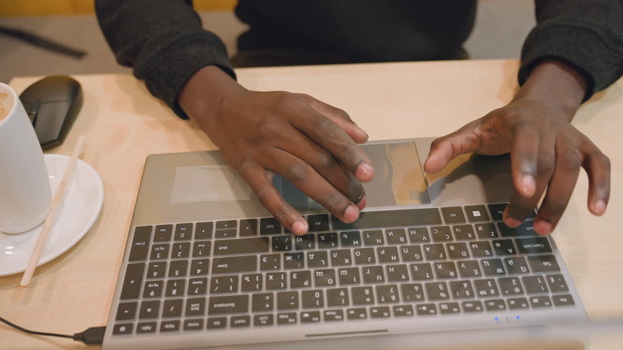 Black hands typing on laptop at desk, entrepreneur preparing presentation slides, cappuccino on saucer, wireless mouse nearby, focused fast typing, modern coworking vibe, bright professional energy