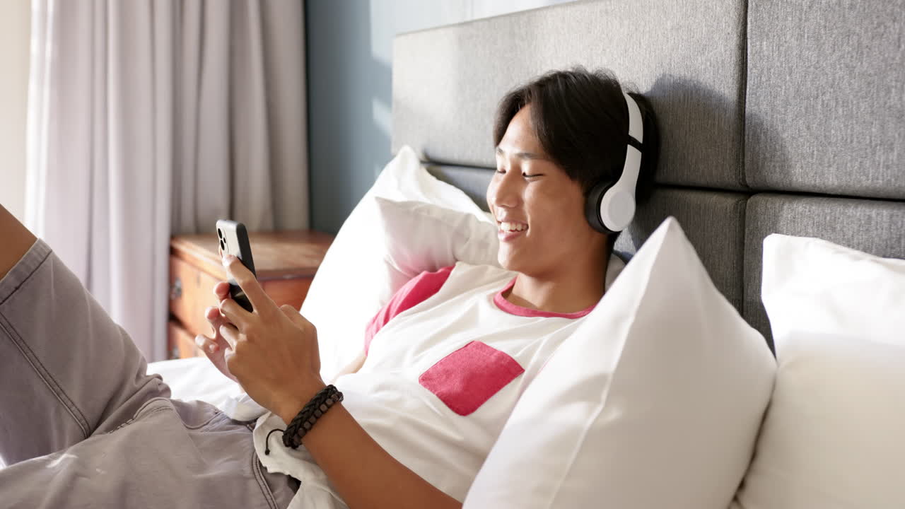Listening to music with headphones, young asian man relaxing on bed using smartphone