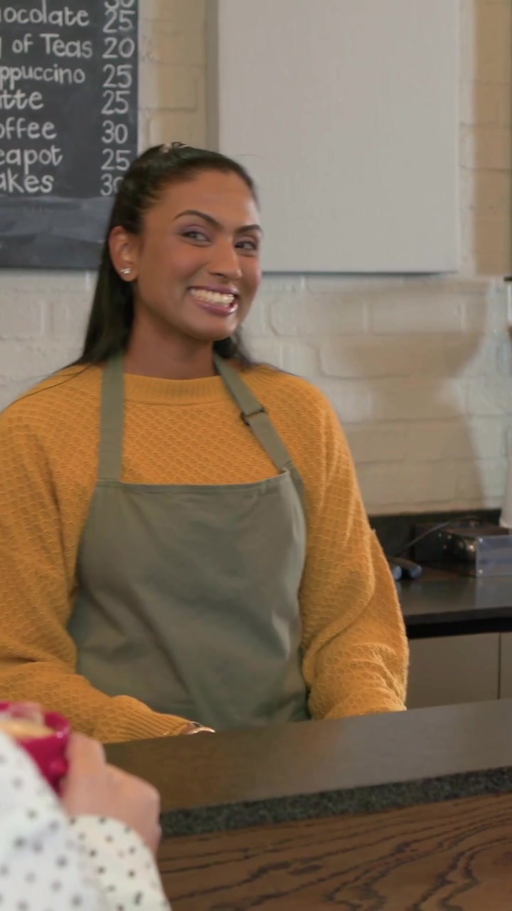 Vertical video: Helping customer holding pink mug, barista wearing apron operating register at cafe