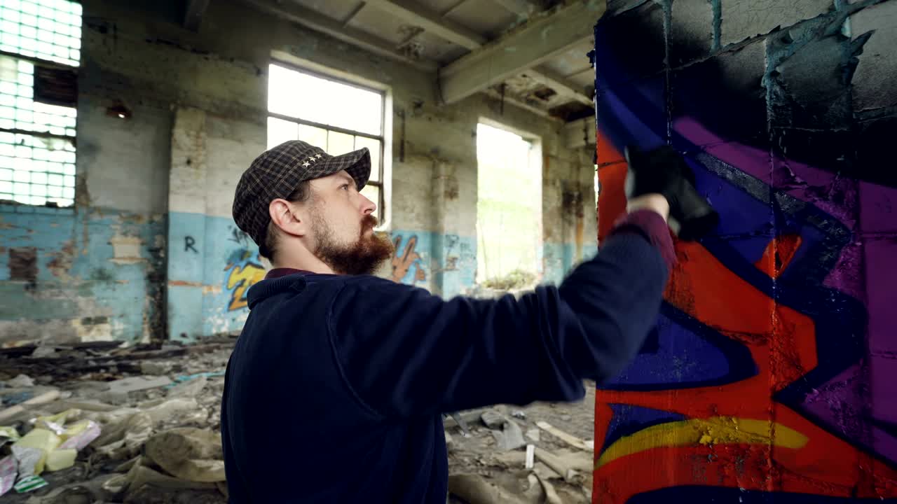 Primo piano di un bell'uomo barbuto, pittore di graffiti, che lavora all'interno di un edificio abbandonato usando vernice spray per creare un'immagine astratta su un pilastro. Creatività e persone.