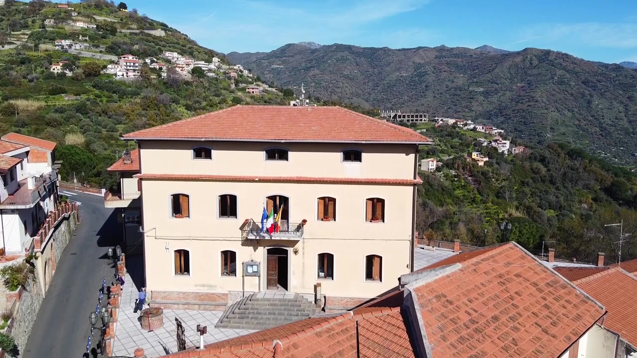 Municipio e strade di Savoca con bandiera siciliana, bandiera italiana sul balcone, esterno di edifici residenziali, balcone con porte e finestre chiuse, valle dei Monti Peloritani aspri