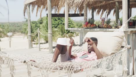 happy african american man lying in hammock putting hat over face on sunny beach, slow motion