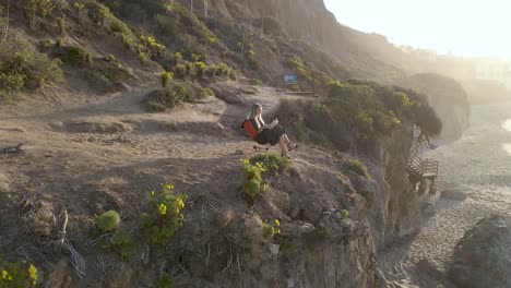 Vista-Aérea-De-Una-Hermosa-Joven-Leyendo-En-Un-Acantilado-Escénico-Al-Atardecer