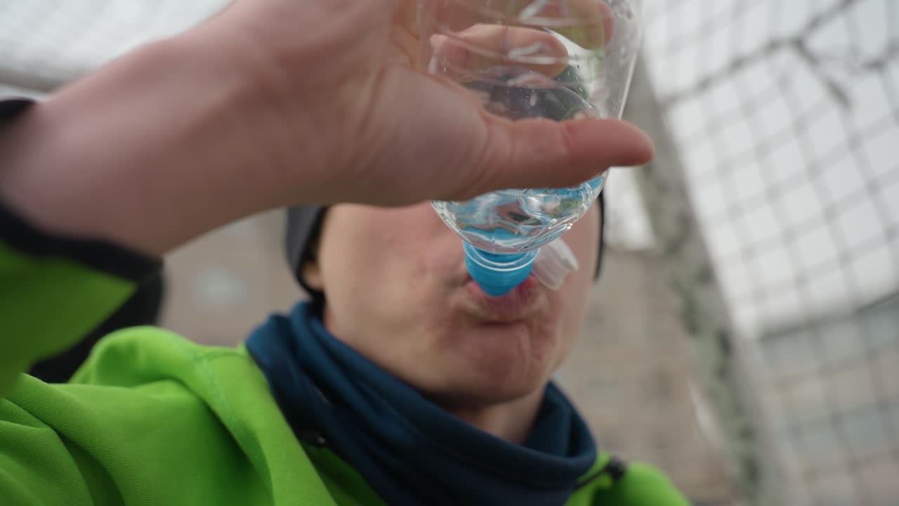 달리는 사람이 물병을 들고 있는 클로즈업 사진입니다. 시점은 손이 플라스틱 병을 입으로 기울이는 것을 보여주며, 녹색 재킷과 검은색 비니, 체인 링크 울타리가 배경으로 보입니다. 차가운 공기 속에서 입김이 보이며, 집중된 표정을 짓고 있습니다.