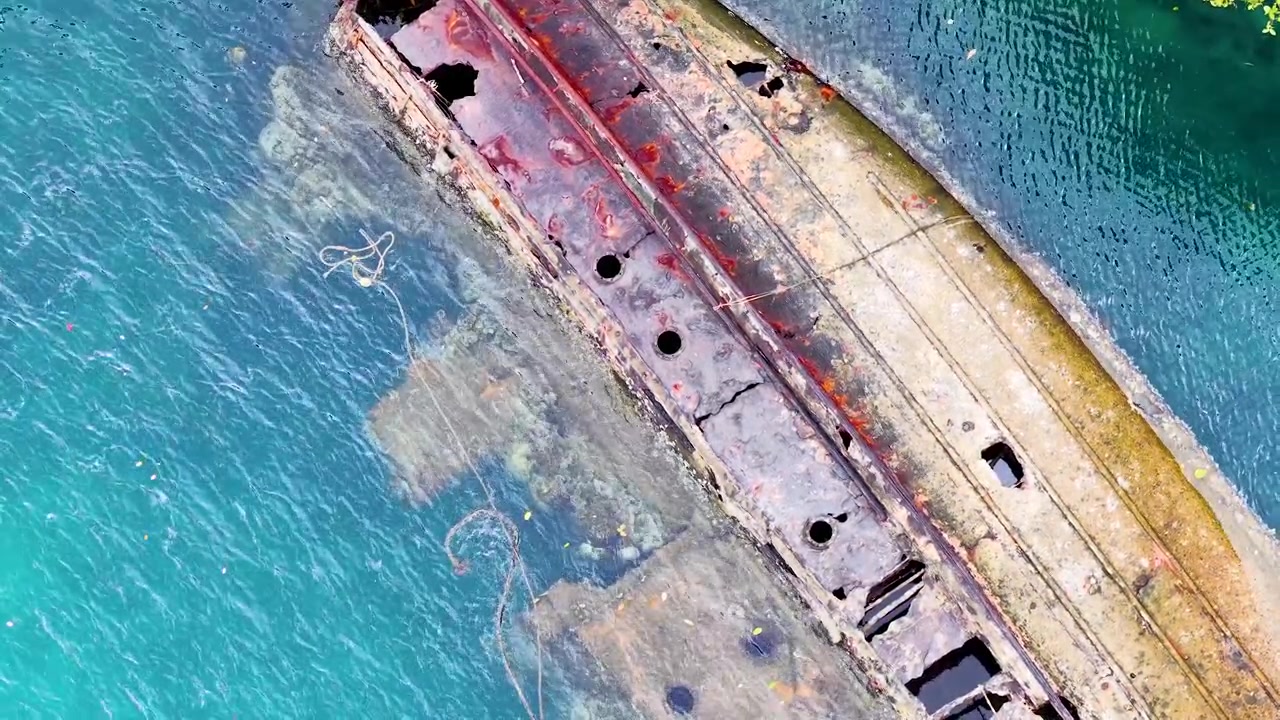 Toma aérea de dron captura un naufragio de metal en descomposición reposando en aguas cristalinas de color turquesa.
