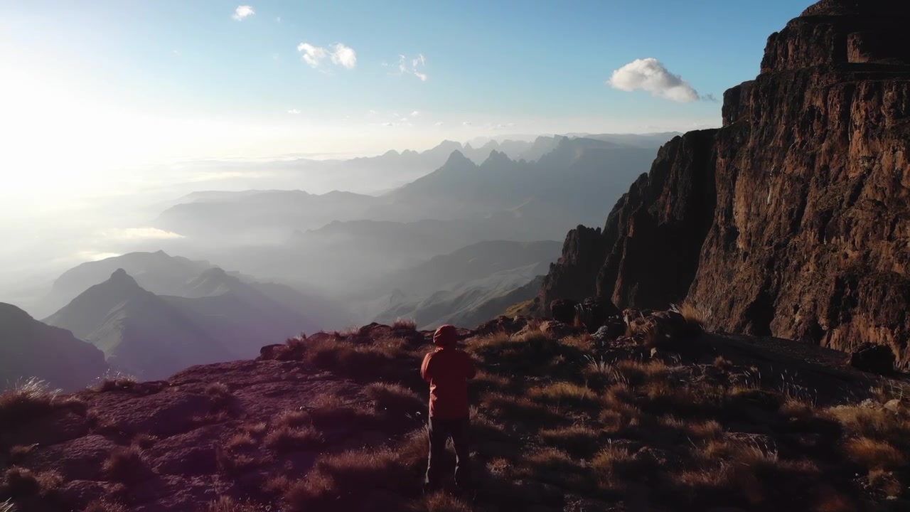 Un escursionista solitario si erge su un altopiano roccioso di montagna che si affaccia su una valle nebbiosa e cime stratificate in lontananza, con luce solare calda, cielo sereno e scogliere imponenti.