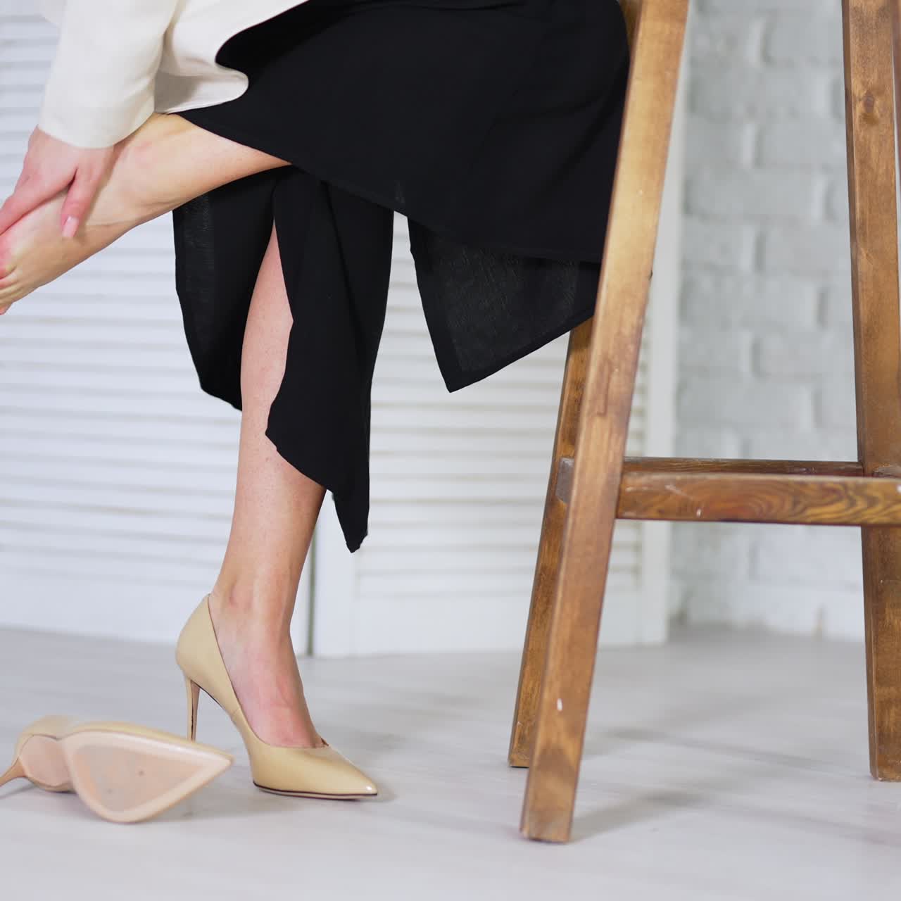 Lady takes off beige high-heeled shoe and feels relief. Lady massages tired foot leaning on a wooden tabouret. White backdrop