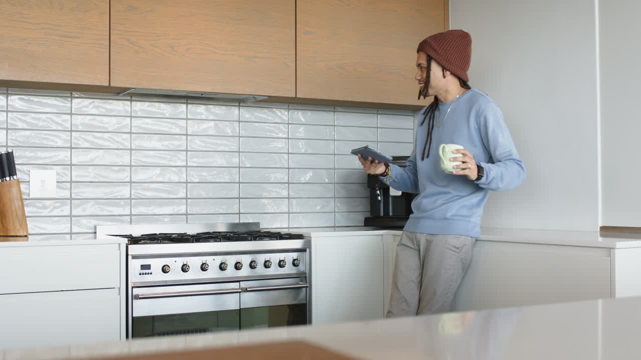 Uomo di mezza età che scorre il tablet e sorseggia caffè al bancone della cucina per seguire una ricetta, con spazio per il testo.