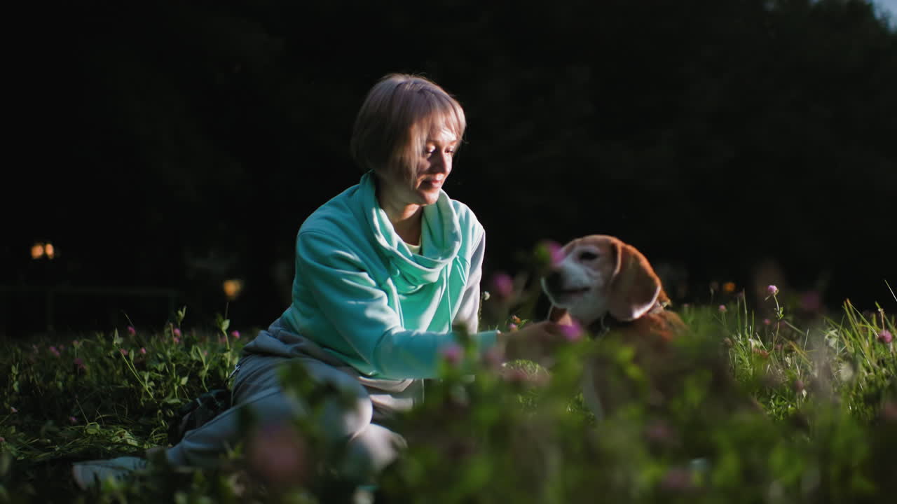 Dog tamer sits on cloth laid over green grass in evening park gently petting her dog under soft light surrounded by blooming flowers creating calm peaceful moment of affection loyalty bonding