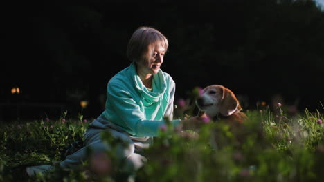 dog tamer sits on cloth laid over green grass in evening park gently petting her dog under soft light surrounded by blooming flowers creating calm peaceful moment of affection loyalty bonding