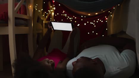 african american daughter and her father watching tablet lying in blanket tent with fairy lights