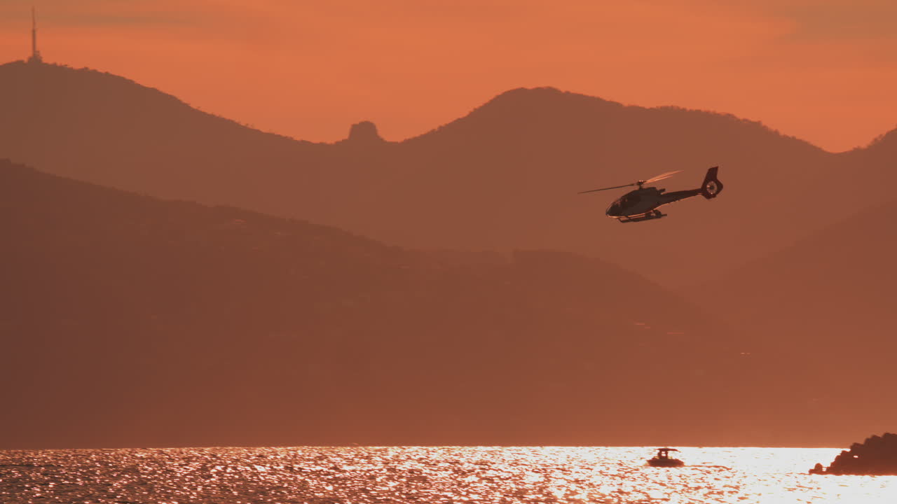 Helicopter flying past a lighthouse with orange sunset sky and distant mountains