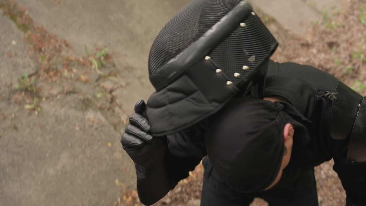 Close up of man in black protective gear adjusting his fencing mask and gloves outdoors on concrete with greenery. subject removes arm guards knee protection helmet pieces while holding