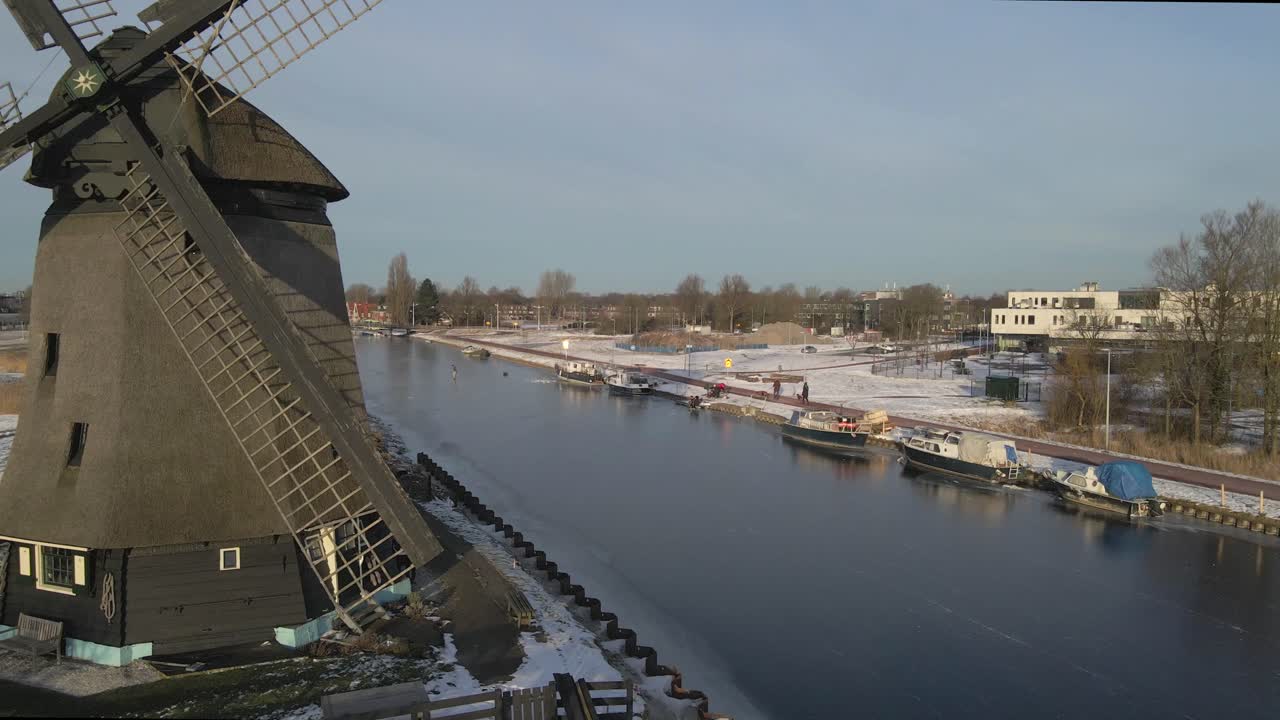 Vista aerea di un mulino a vento che gira con pattinaggio su un fiume ghiacciato all'alba ad Alkmaar, Paesi Bassi, con una splendida luce del sole nascente, un'atmosfera serena e riprese con drone di alta qualità che mostrano paesaggi incantevoli.