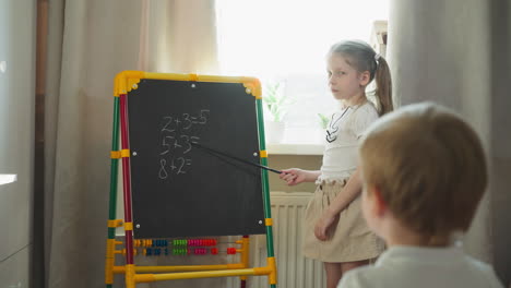 girl shows maths tasks on blackboard to brother at home