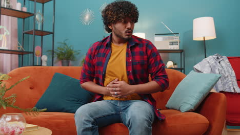 indian man sitting on sofa feeling sudden strong abdominal stomach ache, gastritis problem, diarrhea