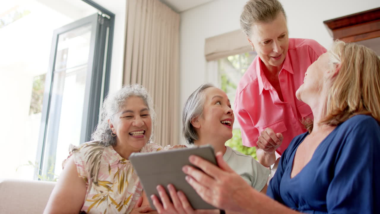 Gruppo eterogeneo di donne anziane che ridono insieme a casa usando un tablet