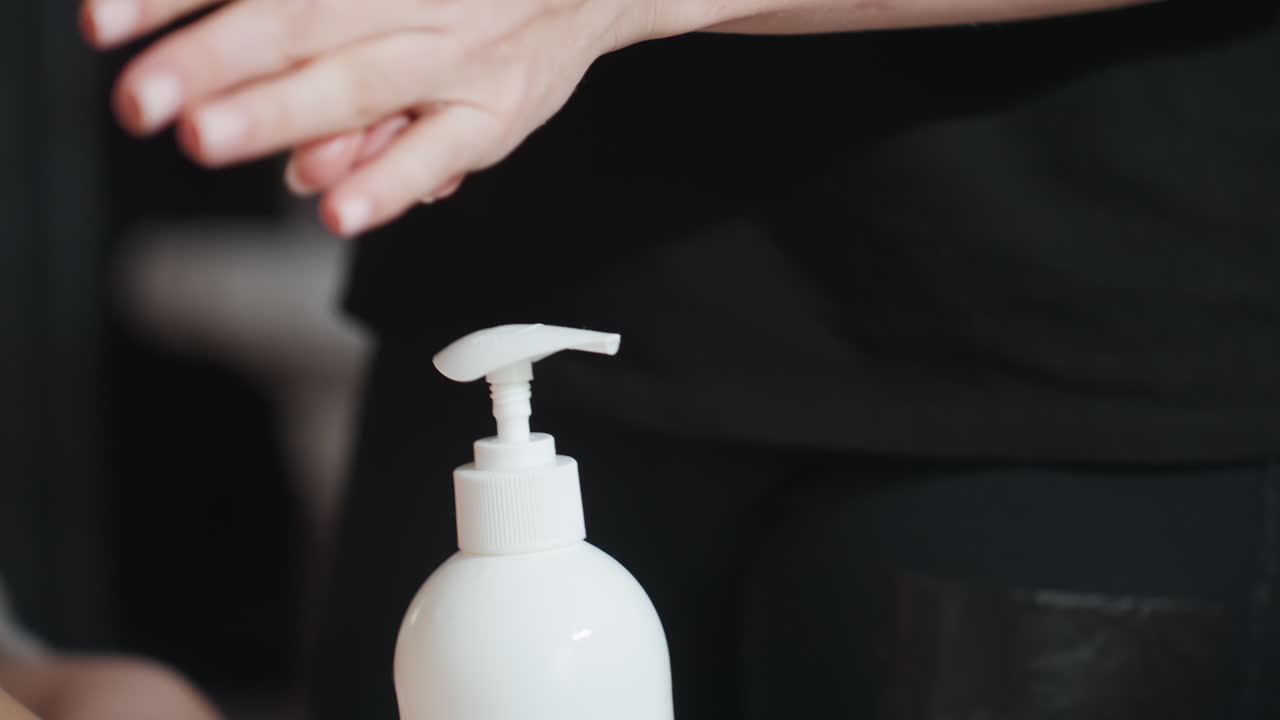 Light skin woman wearing black shirt pumping cream into hand from white bottle, close-up of hands during skincare process, scene highlights hygiene, personal care, and gentle application
